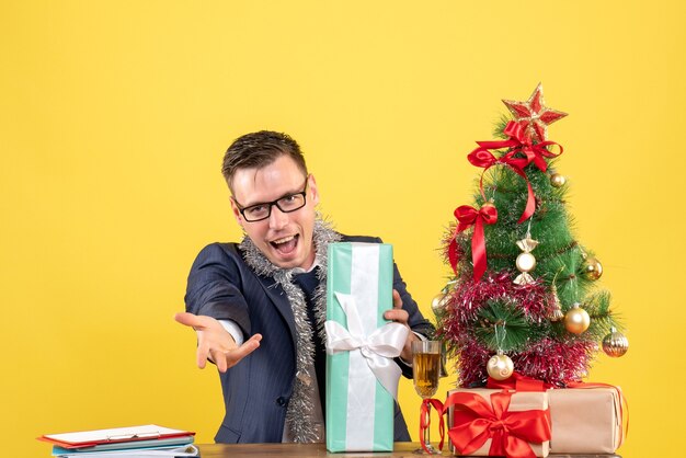 Vorderansicht des glücklichen Mannes, der Hand gibt, die am Tisch nahe Weihnachtsbaum sitzt und auf Gelb präsentiert