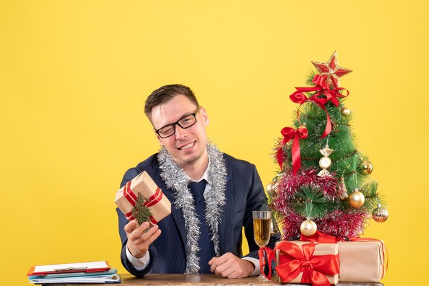 Vorderansicht des glücklichen Mannes mit der Brille, die Geschenk hält, das am Tisch nahe Weihnachtsbaum und Geschenken auf Gelb sitzt