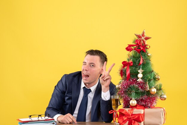 Vorderansicht des gutaussehenden Mannes, der das Siegeszeichen macht, das am Tisch nahe dem Weihnachtsbaum und den Geschenken auf Gelb sitzt