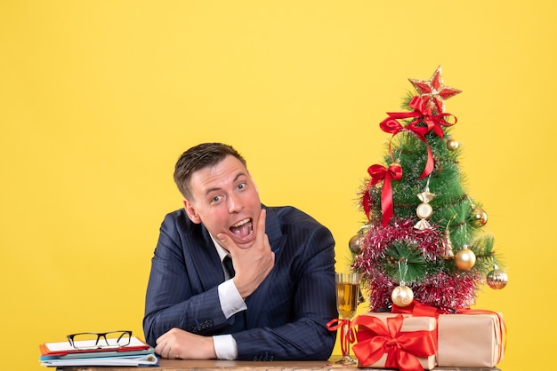 Vorderansicht des gutaussehenden Mannes, der seinen Mund öffnet, der am Tisch nahe Weihnachtsbaum sitzt und auf Gelb präsentiert