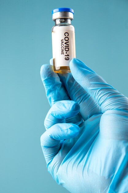 Vorderansicht des Handschuhs, der eine geschlossene Ampulle mit Covid-Impfstoff auf blauem Wellenhintergrund hält