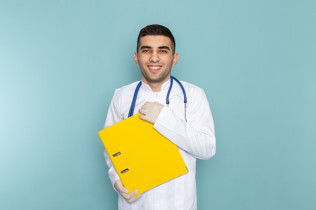 Vorderansicht des jungen männlichen Arztes im weißen Anzug mit blauem Stethoskop, das gelbe Akten hält
