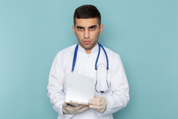 Kostenloses Foto vorderansicht des jungen männlichen arztes im weißen anzug mit blauem stethoskop, das notizblock hält