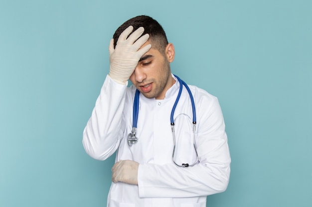 Vorderansicht des jungen männlichen Arztes im weißen Anzug mit blauem Stethoskop, das seinen Kopf hält