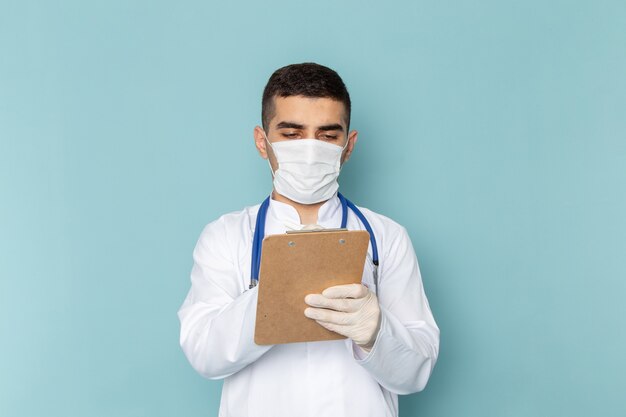 Vorderansicht des jungen männlichen Arztes im weißen Anzug mit blauem Stethoskop, das sterile Maske trägt