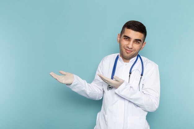 Vorderansicht des jungen männlichen Arztes im weißen Anzug mit dem blauen Stethoskop, das aufwirft und lächelt