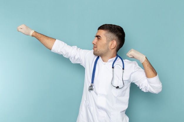 Vorderansicht des jungen männlichen Arztes im weißen Anzug mit dem blauen Stethoskop, das aufwirft