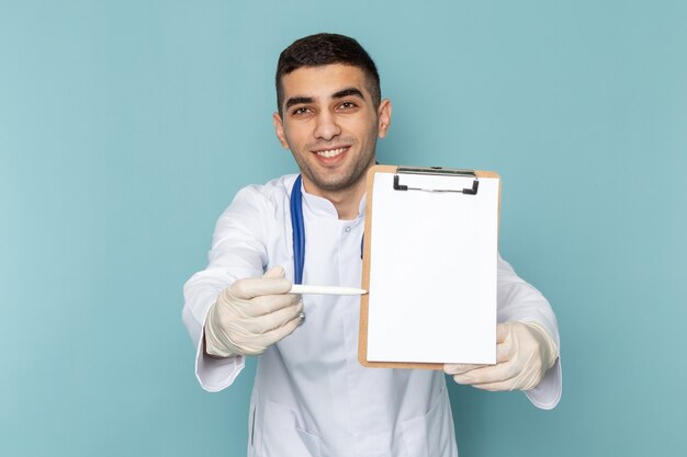Vorderansicht des jungen männlichen Arztes im weißen Anzug mit dem blauen Stethoskop, das Notizen aufschreibt