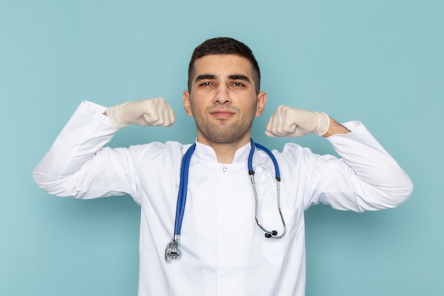 Vorderansicht des jungen männlichen Arztes im weißen Anzug mit dem blauen Stethoskop, das sich biegt