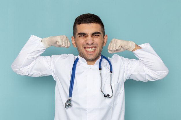 Vorderansicht des jungen männlichen Arztes im weißen Anzug mit dem blauen Stethoskop, das sich biegt