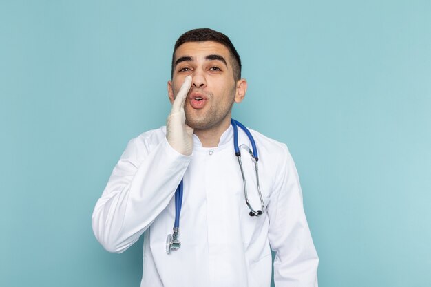 Vorderansicht des jungen männlichen Arztes im weißen Anzug mit Flüstern des blauen Stethoskops