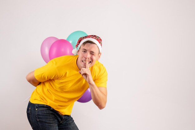 Vorderansicht des jungen Mannes, der bunte Luftballons hinter seinem Rücken auf weißer Wand versteckt