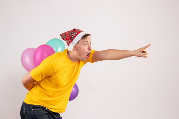 Vorderansicht des jungen Mannes, der bunte Luftballons hinter seinem Rücken auf weißer Wand versteckt