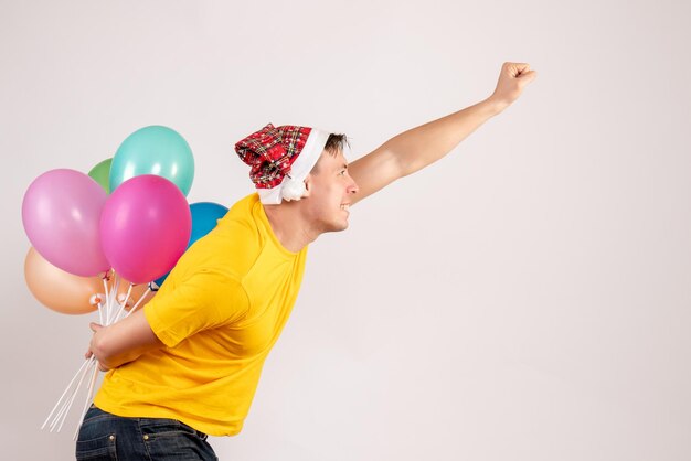 Vorderansicht des jungen Mannes, der bunte Luftballons hinter seinem Rücken auf weißer Wand versteckt