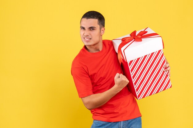 Vorderansicht des jungen Mannes, der Geschenk im Kasten auf gelber Wand hält