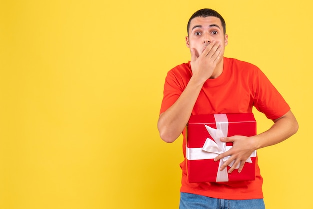 Vorderansicht des jungen Mannes, der Geschenk schockiert auf gelber Wand hält