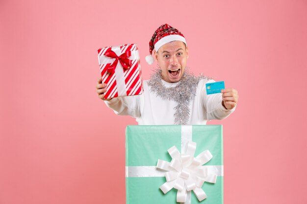 Vorderansicht des jungen Mannes, der Geschenk und Bankkarte an der rosa Wand hält