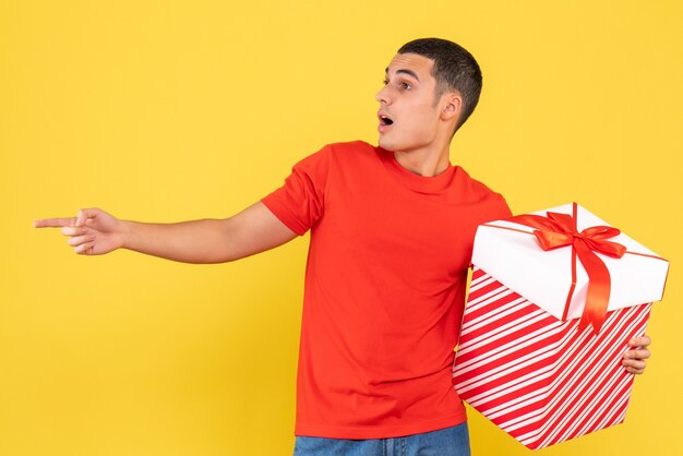 Vorderansicht des jungen Mannes, der großes Weihnachtsgeschenk auf gelber Wand hält