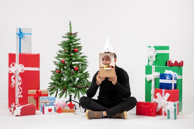 Vorderansicht des jungen Mannes, der um Geschenke auf einer weißen Wand sitzt