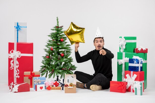 Vorderansicht des jungen Mannes, der um Geschenke sitzt und Goldsternfigur auf weißer Wand hält