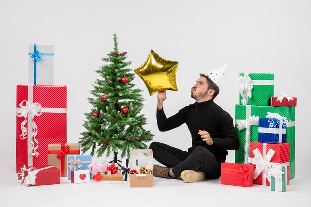 Vorderansicht des jungen Mannes, der um Geschenke sitzt und Goldsternfigur auf weißer Wand hält