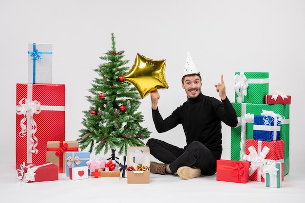 Vorderansicht des jungen Mannes, der um Geschenke sitzt und Goldsternfigur auf weißer Wand hält