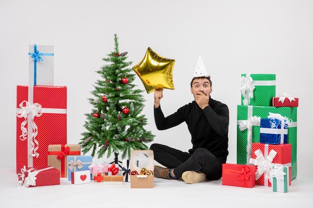 Vorderansicht des jungen Mannes, der um Geschenke sitzt und Goldsternfigur auf weißer Wand hält