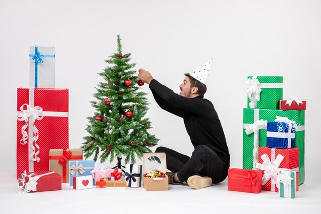 Vorderansicht des jungen Mannes, der um Urlaubsgeschenke sitzt, verziert kleinen Baum auf einer weißen Wand