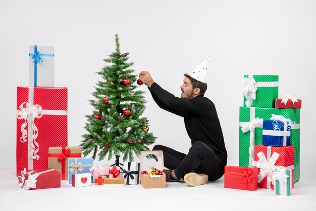 Vorderansicht des jungen Mannes, der um Urlaubsgeschenke sitzt, verziert kleinen Baum auf einer weißen Wand