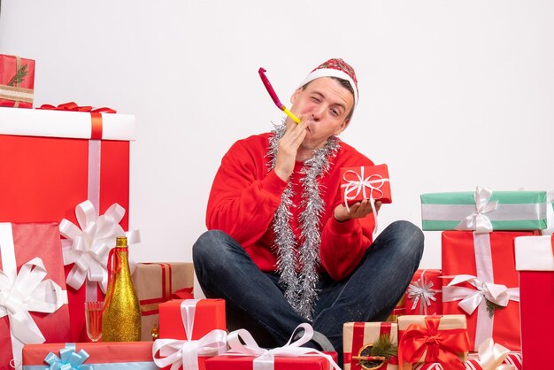 Vorderansicht des jungen Mannes, der um Weihnachtsgeschenke auf weißer Wand sitzt