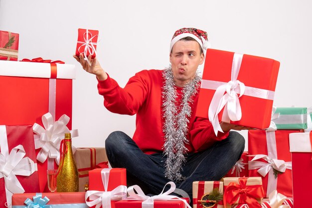 Vorderansicht des jungen Mannes, der um Weihnachtsgeschenke auf weißer Wand sitzt