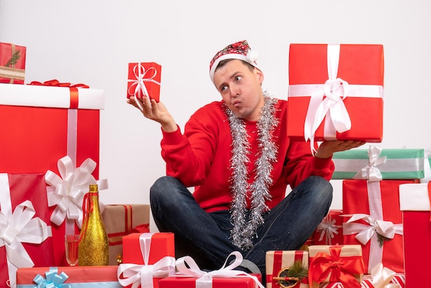 Vorderansicht des jungen Mannes, der um Weihnachtsgeschenke auf weißer Wand sitzt