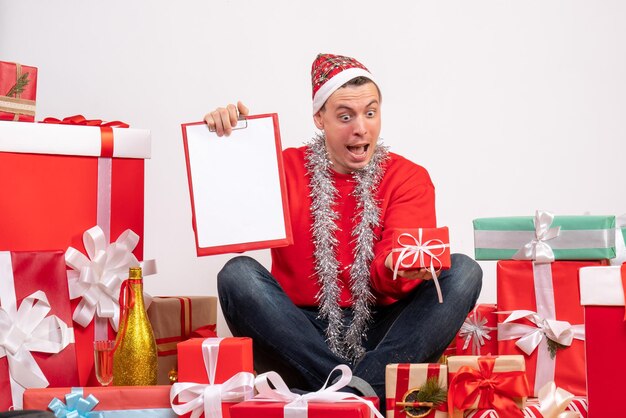 Vorderansicht des jungen Mannes, der um Weihnachtsgeschenke herum sitzt, mit Hinweis an der weißen Wand