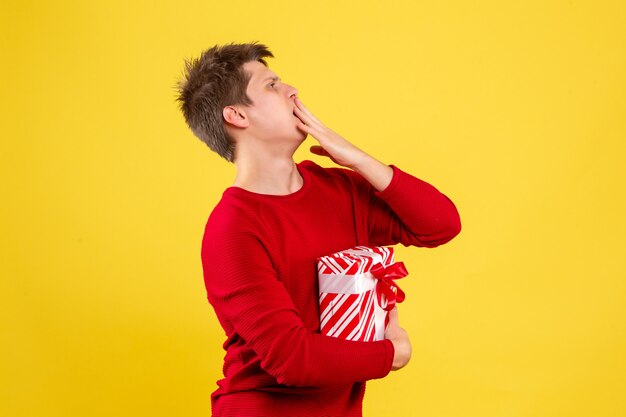 Vorderansicht des jungen Mannes, der Weihnachtsgeschenk auf gelber Wand hält