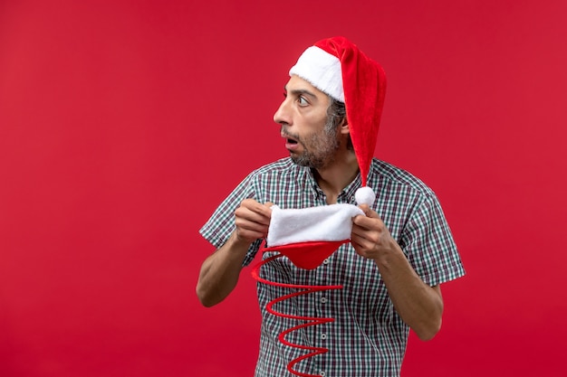 Vorderansicht des jungen Mannes, der Weihnachtsspielzeugmütze an der roten Wand hält