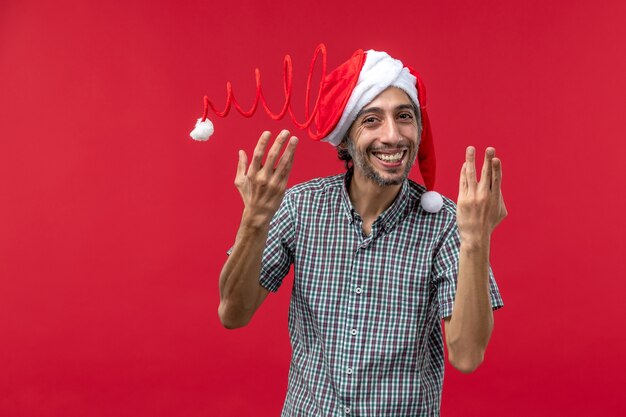 Vorderansicht des jungen Mannes, der Weihnachtsspielzeugmütze an der roten Wand trägt