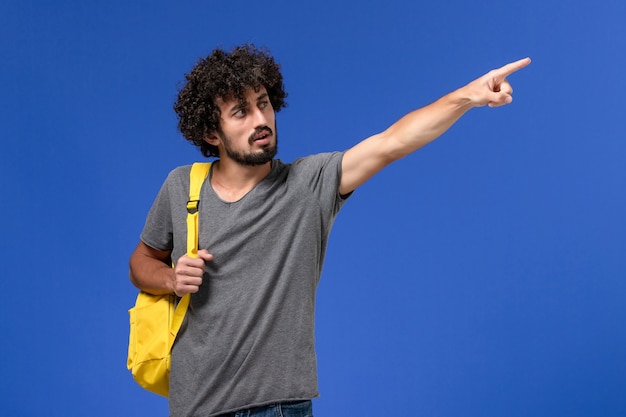 Vorderansicht des jungen Mannes im grauen T-Shirt, das gelben Rucksack an der blauen Wand trägt