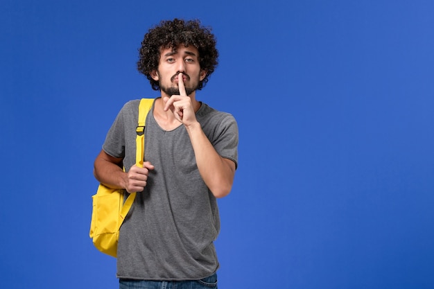 Vorderansicht des jungen Mannes im grauen T-Shirt, das gelben Rucksack an der blauen Wand trägt