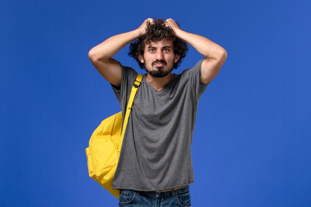 Vorderansicht des jungen Mannes im grauen T-Shirt, das gelben Rucksack auf der hellblauen Wand trägt