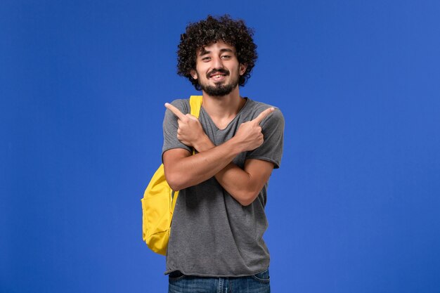 Vorderansicht des jungen Mannes im grauen T-Shirt, das gelben Rucksack lächelnd auf der blauen Wand posierend trägt