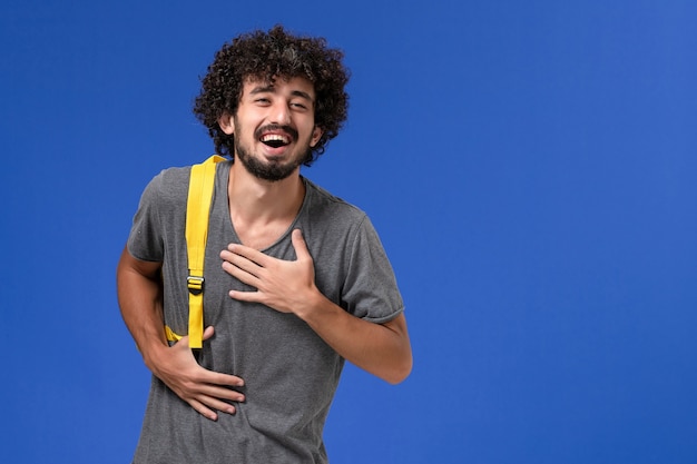 Vorderansicht des jungen Mannes im grauen T-Shirt, das gelben Rucksack trägt, der an der blauen Wand lacht