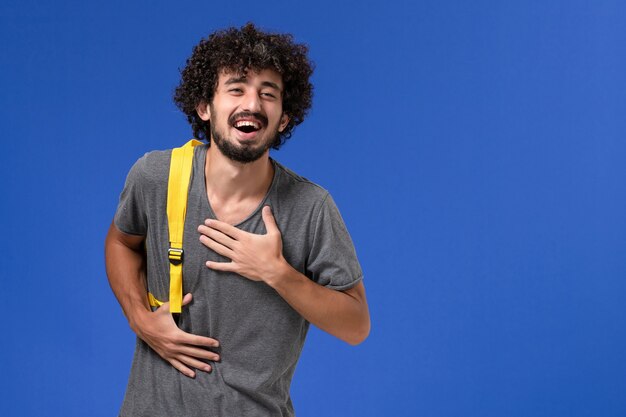 Vorderansicht des jungen Mannes im grauen T-Shirt, das gelben Rucksack trägt, der an der blauen Wand lacht