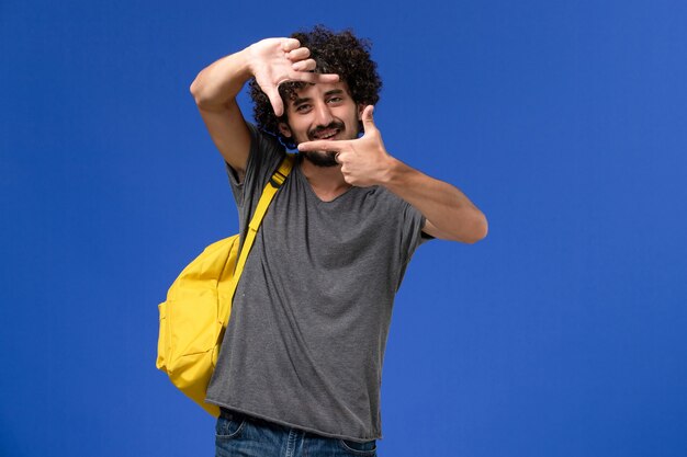 Vorderansicht des jungen Mannes im grauen T-Shirt, das gelben Rucksack trägt, der auf blauer Wand aufwirft