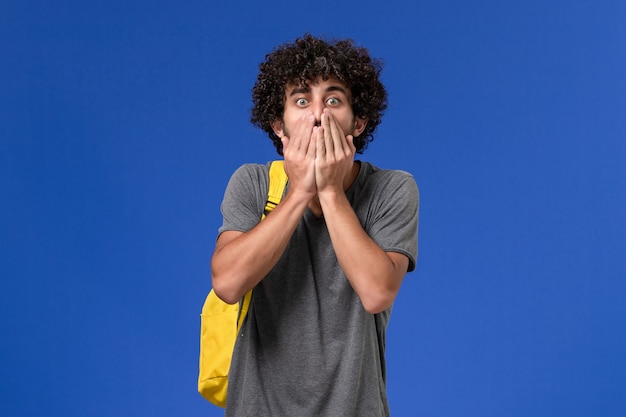 Vorderansicht des jungen Mannes im grauen T-Shirt, das gelben Rucksack trägt, der mit schockiertem Ausdruck auf der blauen Wand aufwirft