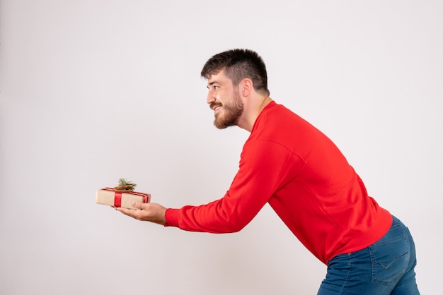 Vorderansicht des jungen Mannes im roten Hemd, das Weihnachtsgeschenk auf einer weißen Wand gibt