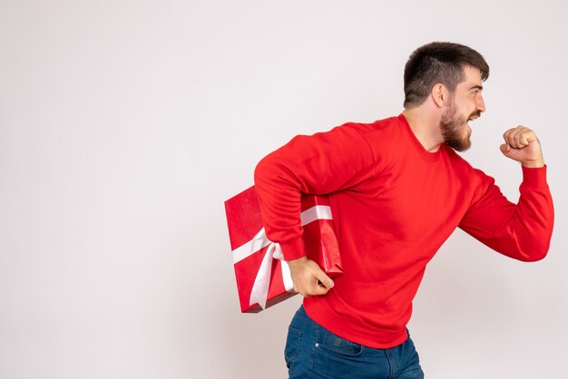 Vorderansicht des jungen Mannes im roten Hemd, das Weihnachtsgeschenk auf weißer Wand hält