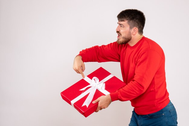 Vorderansicht des jungen Mannes im roten Hemd, das Weihnachtsgeschenk auf weißer Wand hält