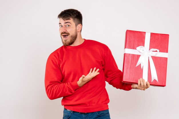 Vorderansicht des jungen Mannes im roten Hemd, das Weihnachtsgeschenk auf weißer Wand hält