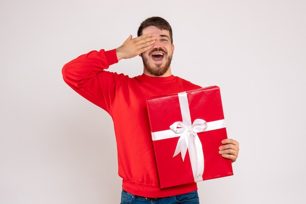 Vorderansicht des jungen Mannes im roten Hemd, das Weihnachtsgeschenk auf weißer Wand hält