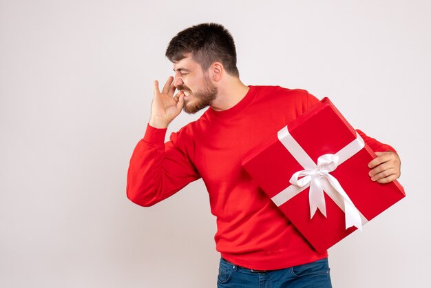 Vorderansicht des jungen Mannes im roten Hemd, das Weihnachtsgeschenk auf weißer Wand hält